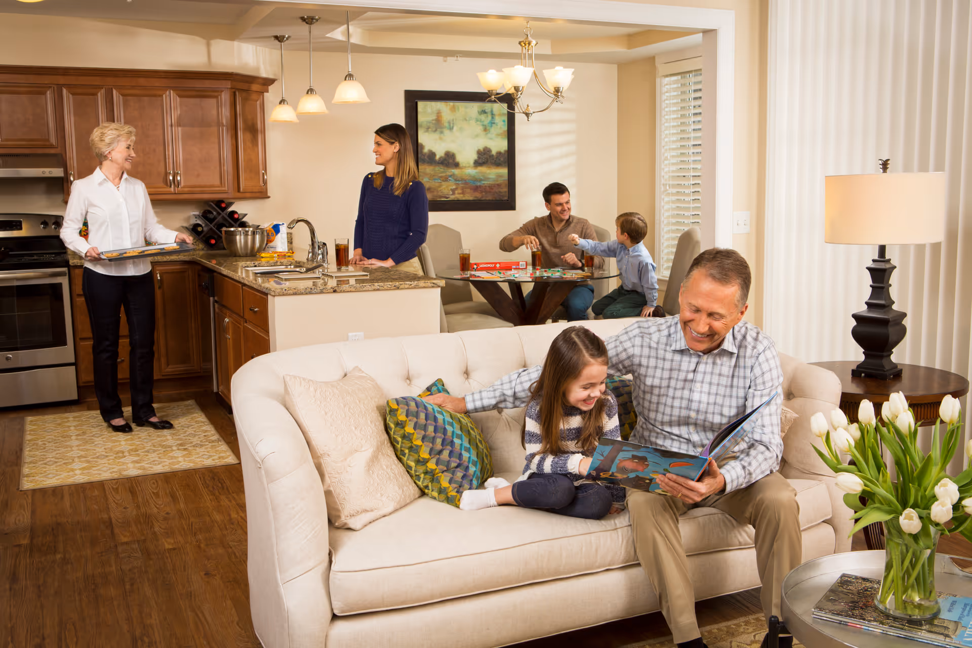 A multi-generational family scene inside a cozy home. In the foreground, an older man sits on a beige couch reading a book to a young girl. Behind them, a woman stands in a kitchen area holding a tray, while another woman leans on the kitchen counter. In the background, a man and a young boy play a board game at a dining table. The room is warmly lit with wooden floors, a lamp on a side table, and a vase of white tulips on a coffee table.