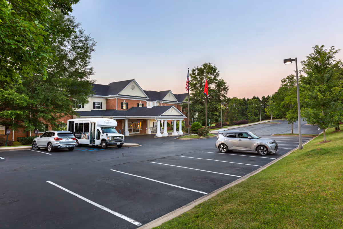 Front entrance of a two-story brick senior living facility with a covered porte-cochère, parked cars and a shuttle in the parking lot and flagpoles in front.