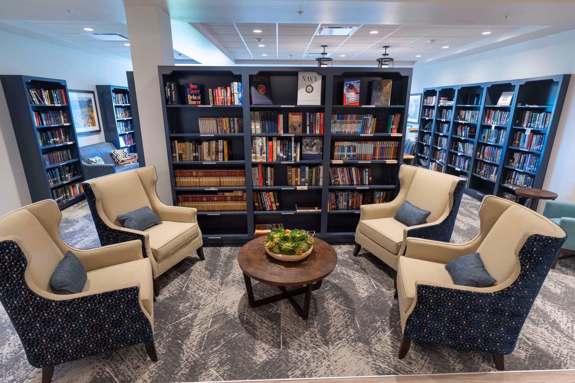 A cozy library room in a senior living facility with four beige and blue armchairs arranged around a round wooden coffee table with a plant centerpiece. Behind the seating area are tall black bookshelves filled with books. Additional bookshelves and seating areas are visible in the background.