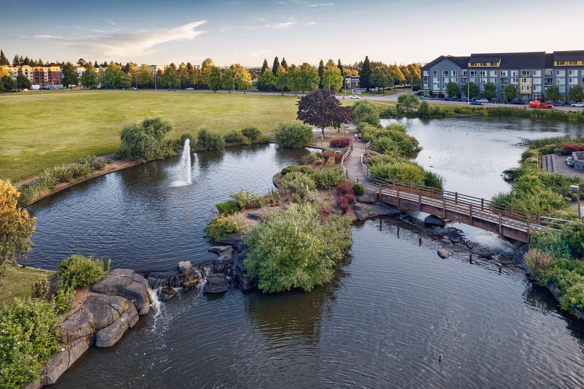 A scenic outdoor park area with a large pond featuring a water fountain and small waterfall. There is a wooden footbridge crossing part of the pond, surrounded by lush greenery and trees. In the background, there are residential buildings and a grassy field under a partly cloudy sky during sunset.