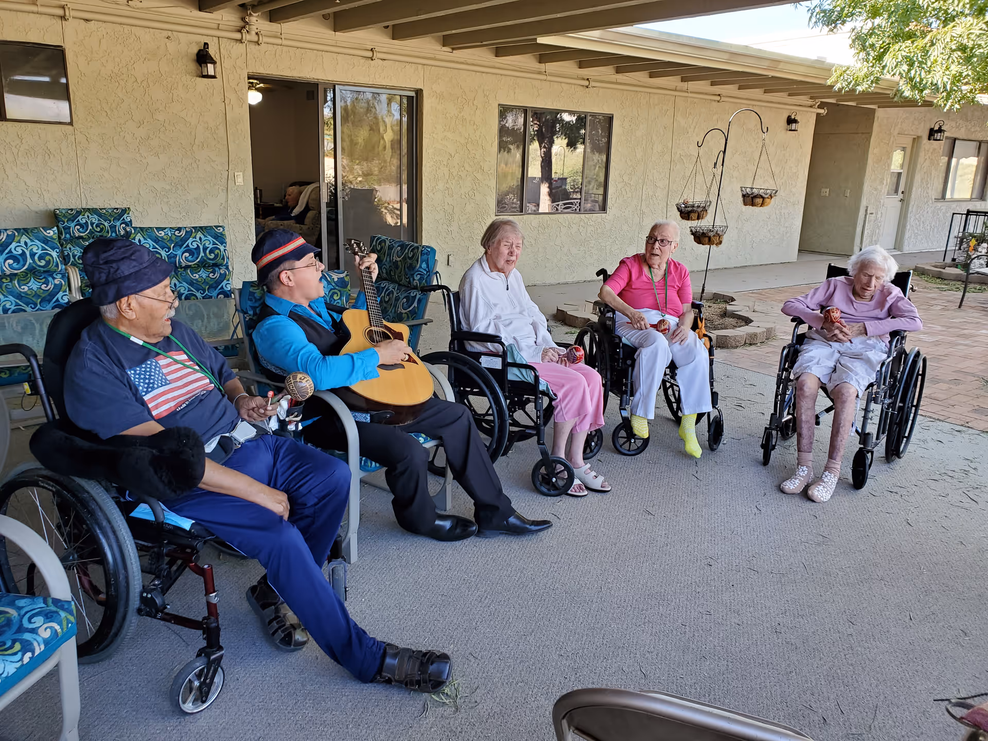 Five elderly individuals sitting outside on a covered patio area, four in wheelchairs and one seated on a chair, with one person playing a guitar. The patio has patterned blue cushions on chairs, hanging planters, and a beige stucco wall with windows and a sliding glass door in the background.