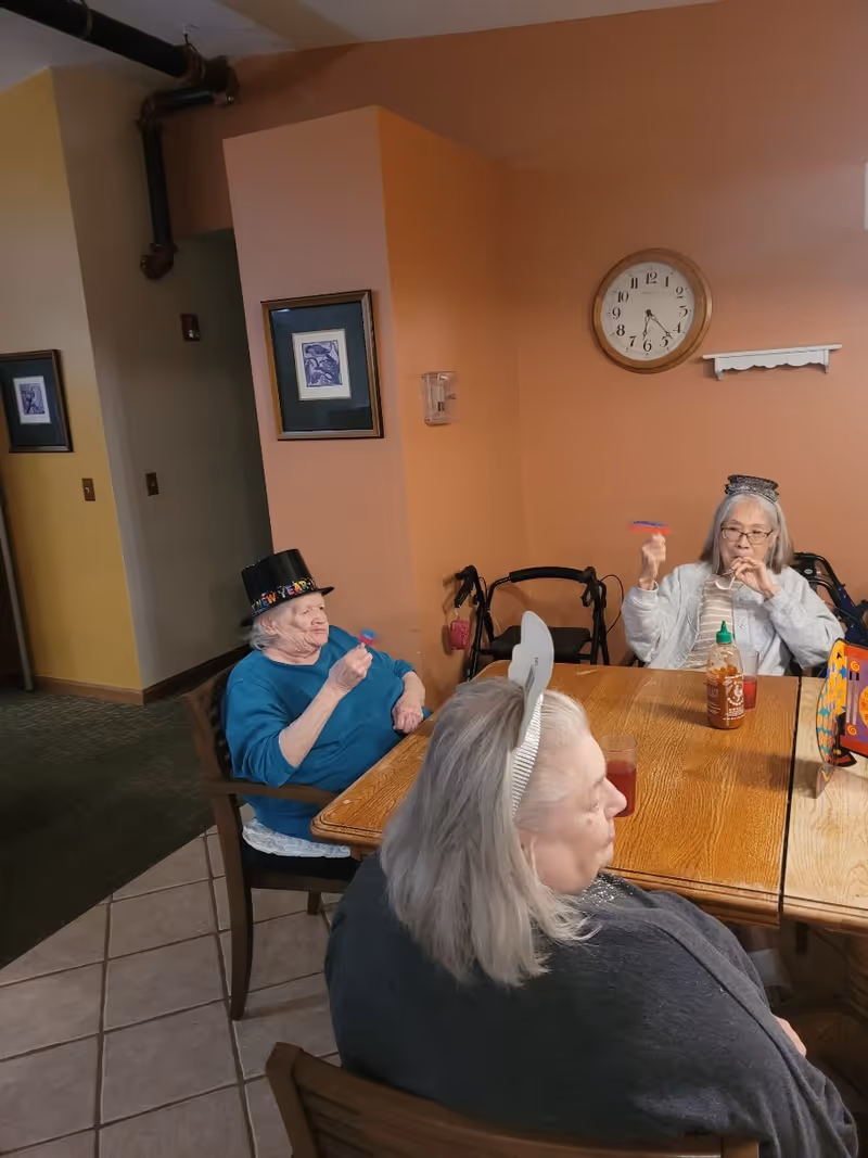 Three elderly women sitting around a wooden table in a room with peach-colored walls. Two women are wearing festive hats and blowing party blowers. There is a clock on the wall showing the time as 6:25, framed pictures, and a bottle of hot sauce on the table.