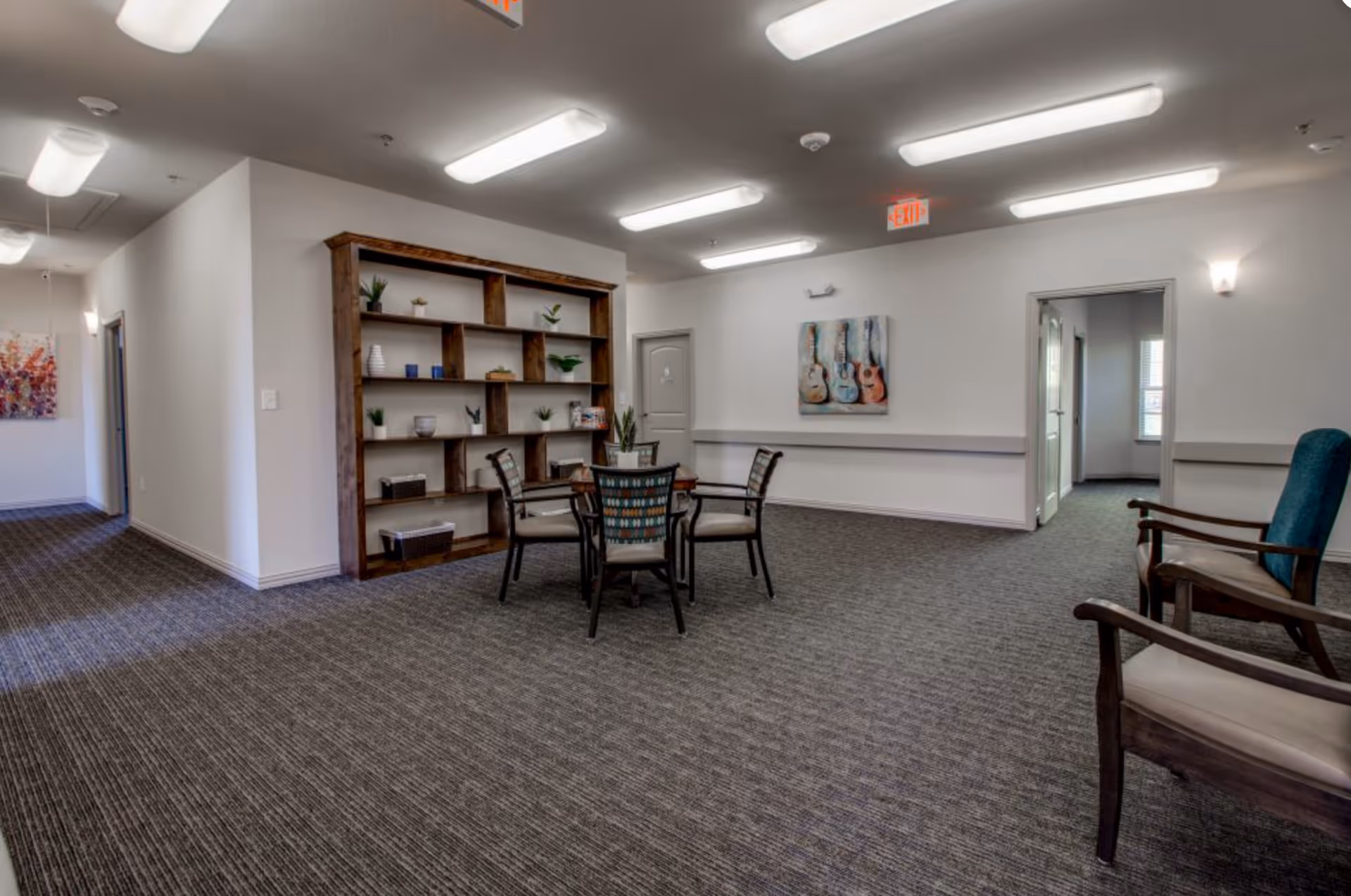 A spacious common lounge with a central table and chairs, shelving, and additional seating under ceiling lights.