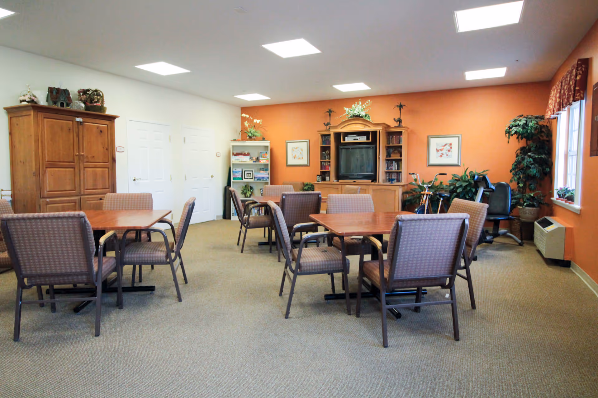 A common room with several tables and chairs arranged for group seating. The room has an orange accent wall with a wooden entertainment center holding a TV and various decorative items. There are plants, framed pictures, and a bookshelf with games and books. The room is well-lit with ceiling lights and has a window with patterned curtains on the right side.
