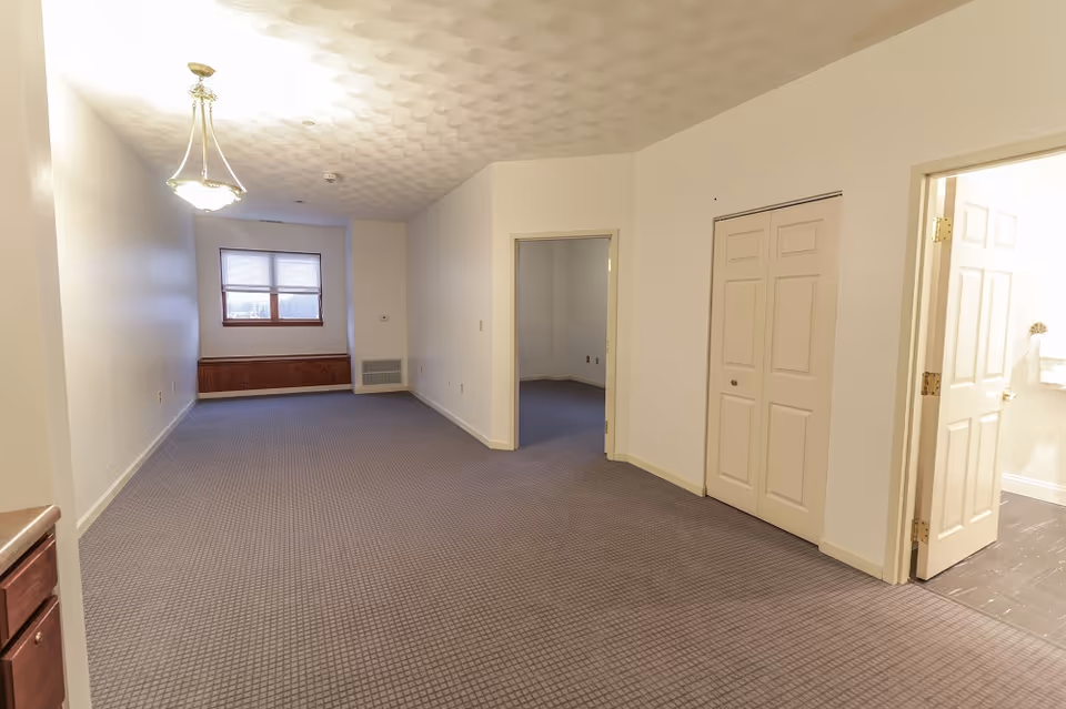 Empty interior room with beige walls and carpeted floor, featuring a window with blinds at the far end, a ceiling light fixture, an open doorway leading to another room, a closed closet with double doors, and an open door revealing a bathroom with tiled floor.