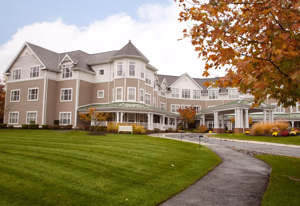Exterior view of a large, multi-story senior living facility with beige siding and white trim. The building features multiple windows, a covered entrance with white columns, and a well-maintained green lawn with a curved walkway. Trees with autumn-colored leaves are visible on the right side.
