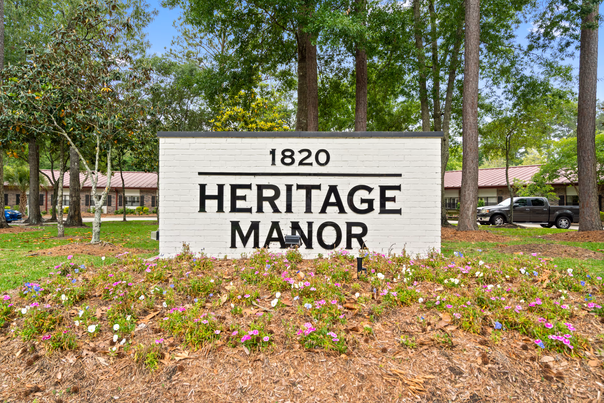White brick sign reading "Heritage Manor" in a landscaped bed of flowers with trees and the facility building behind.