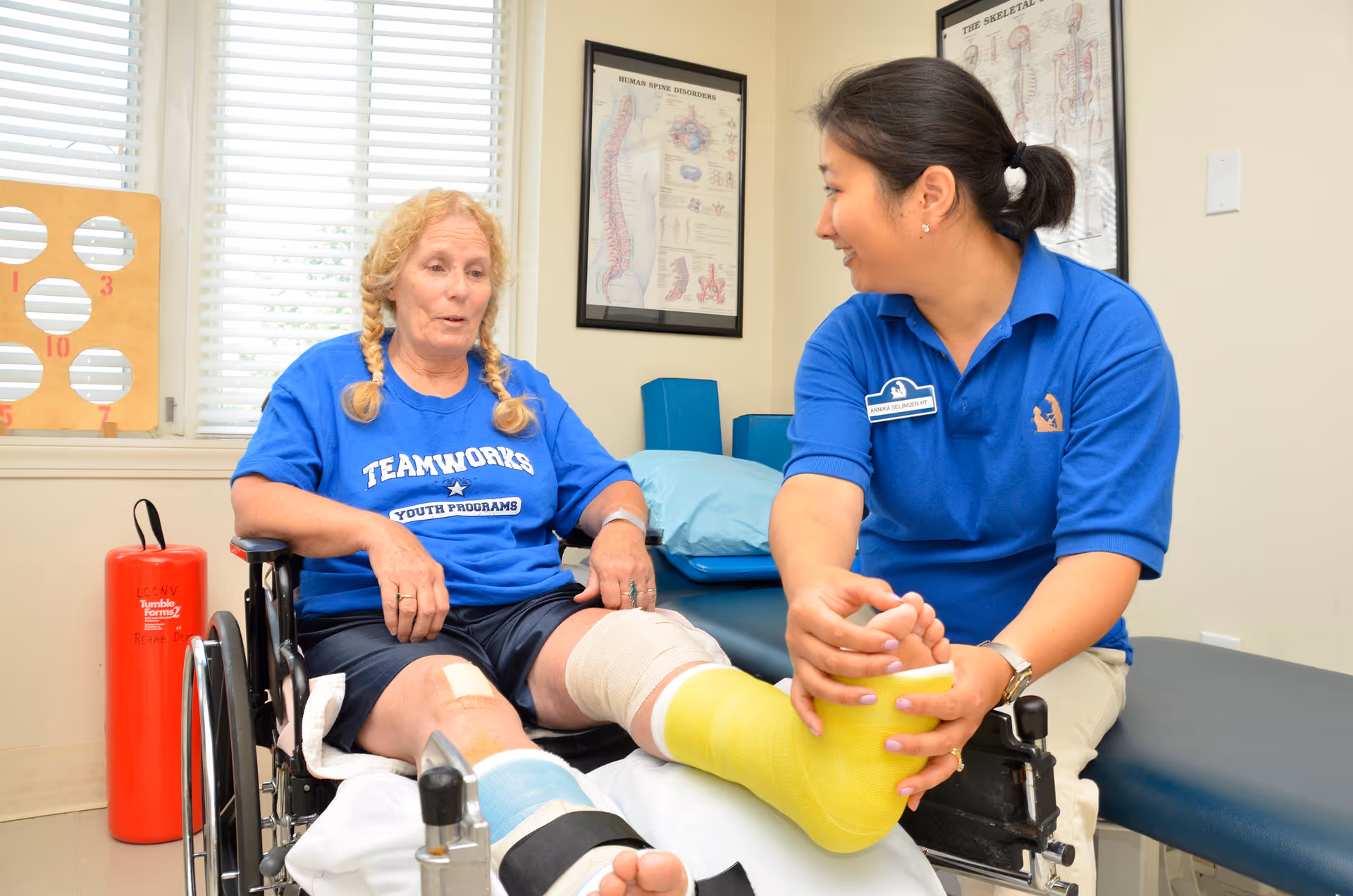 A female healthcare professional in a blue polo shirt is assisting an elderly woman in a wheelchair with a yellow cast on her leg. They are in a medical or physical therapy room with anatomical charts on the wall and a red fire extinguisher nearby.