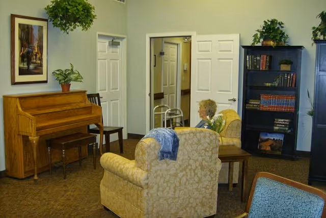 A cozy living room in a memory care facility with two beige patterned armchairs, a wooden piano with a bench, a black bookshelf filled with books and plants, a framed painting on the wall, and a seated elderly woman facing away. The room has beige walls, carpeted floor, and white doors, one of which is open showing a hallway with a walker.
