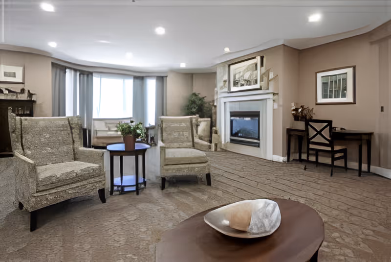 A cozy living room with two patterned armchairs around a small round table with a potted plant. There is a fireplace with a framed picture above it, a desk with a chair, and a large window with curtains letting in natural light. The room has beige walls and carpeted flooring.