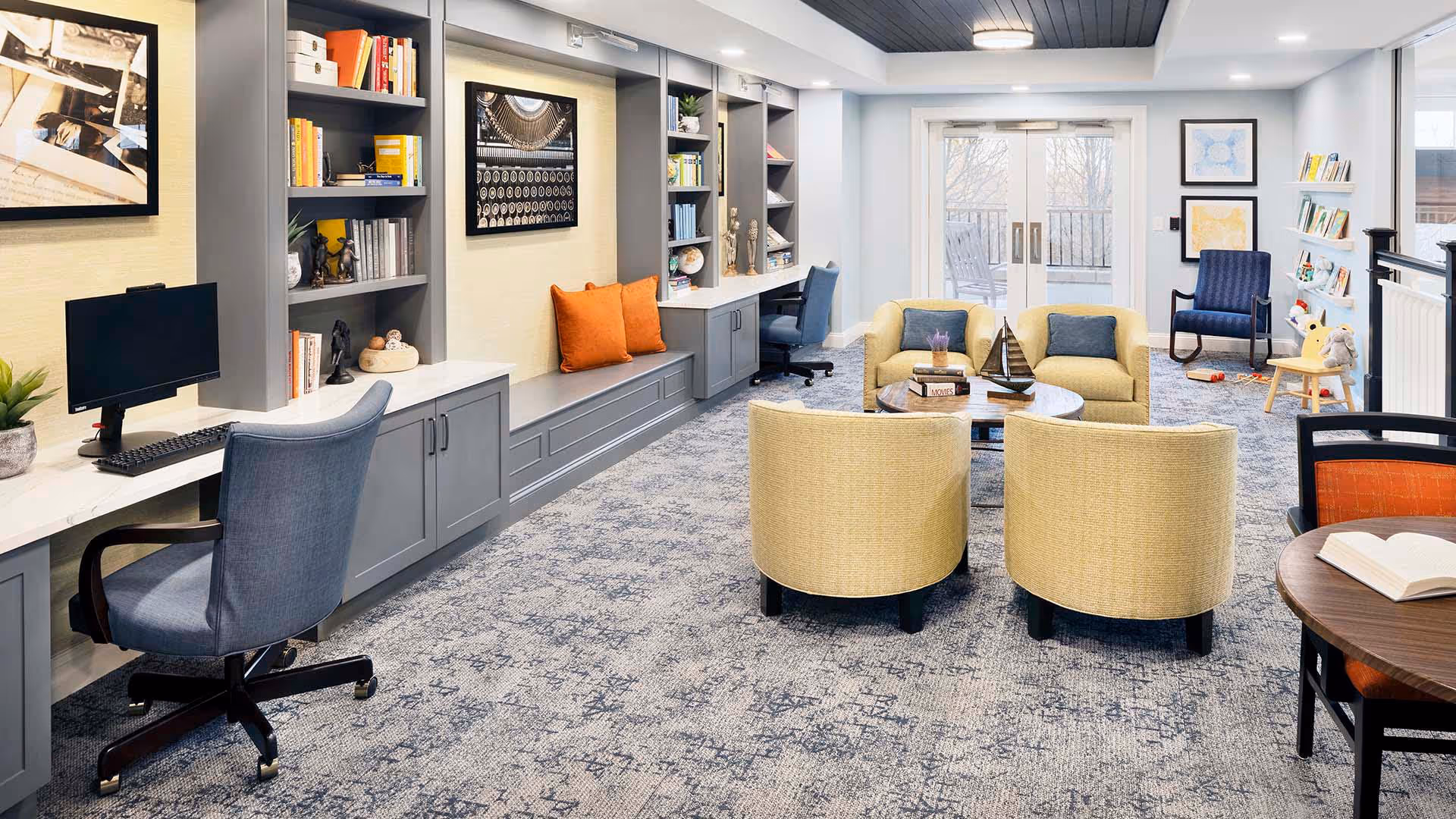 A bright and spacious common area with a seating arrangement including four yellow armchairs around a round coffee table. The room features built-in gray shelving and cabinetry along one wall, decorated with books, plants, and art pieces. There is a desk with a computer and an office chair on the left side. Large windows and glass doors at the far end let in natural light, and a blue armchair and children's toys are visible near the windows. The room has a patterned carpet and a modern ceiling light.
