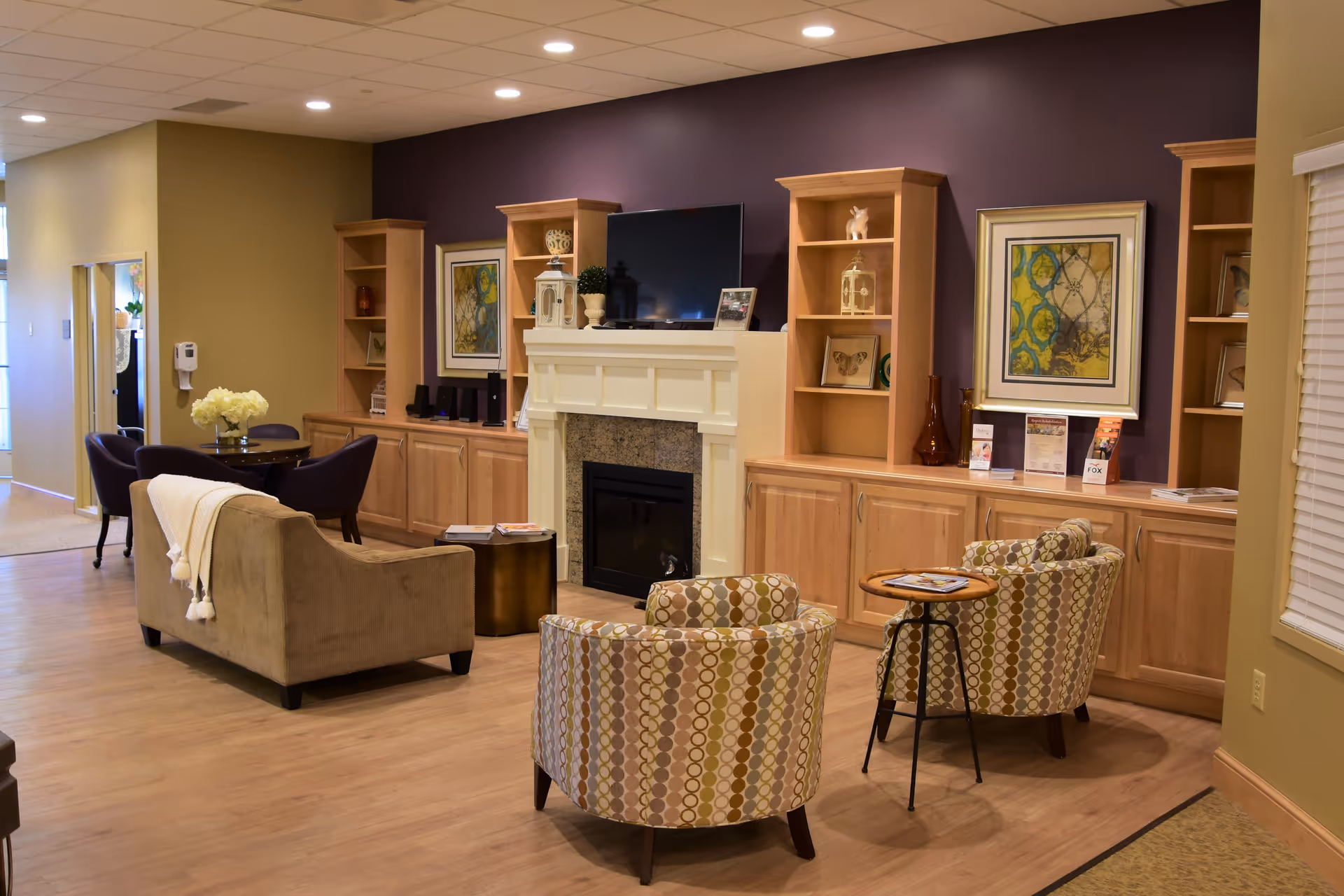 A cozy living room area in an assisted living facility featuring a beige sofa with a white throw, two patterned armchairs, a small round wooden side table, and a fireplace with a mounted flat-screen TV above it. The room has built-in wooden shelves and cabinets along a purple accent wall, decorated with framed artwork and various decorative items. There is also a small round dining table with four chairs and a vase of flowers in the background.