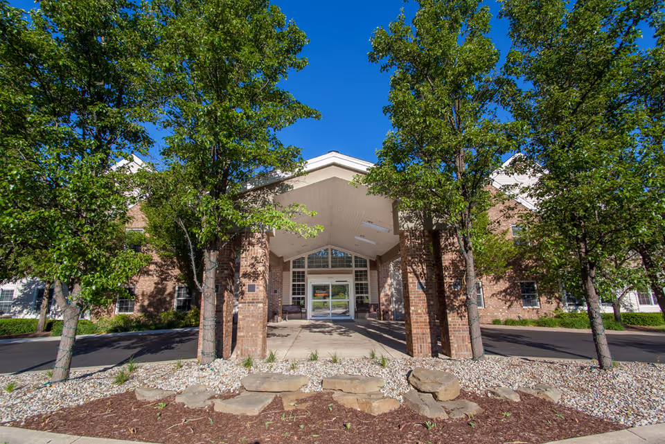 Front entrance of a brick building with a covered driveway supported by brick pillars, surrounded by green trees and landscaping with rocks and mulch under a clear blue sky.