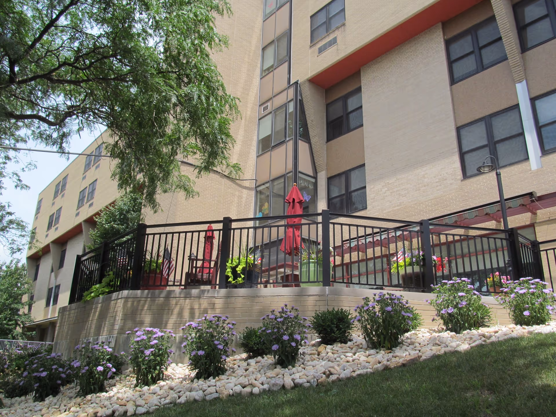 Raised outdoor patio with railing, potted plants and umbrellas in front of a multi-story brick nursing facility.