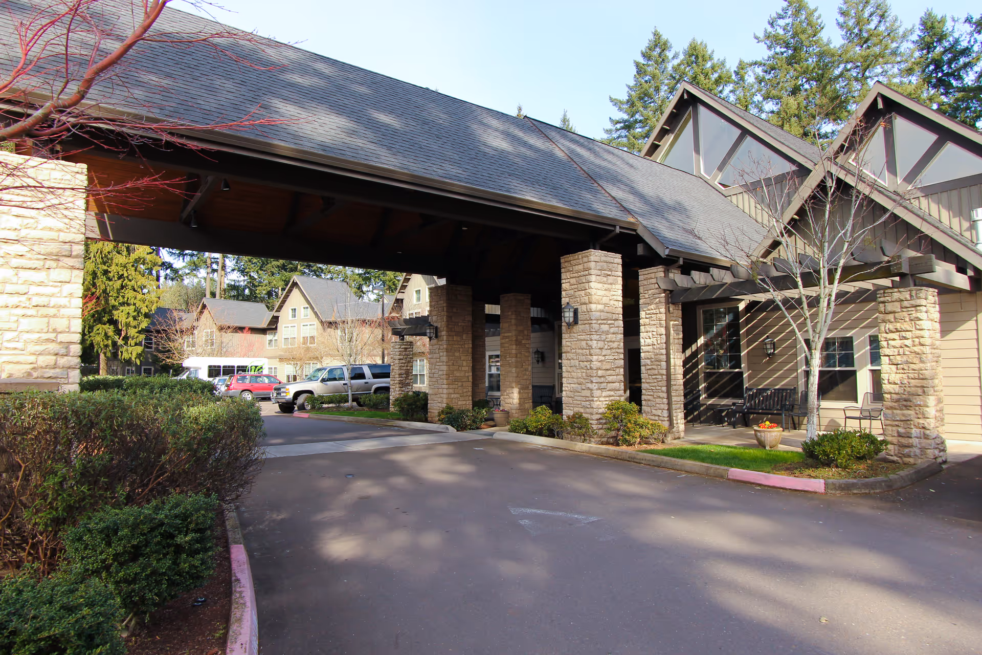 Exterior view of a senior living facility entrance with a covered driveway supported by stone pillars. There are bushes and trees around the driveway, several parked vehicles, and residential-style buildings in the background under a clear sky.