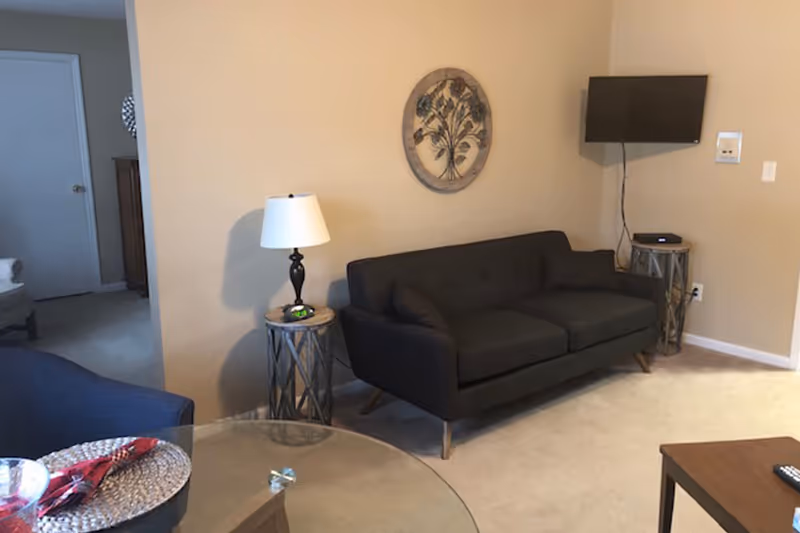 A living room with a dark gray sofa against a beige wall, a small round side table with a lamp and a clock, a wall-mounted flat screen TV, and a glass-top table with a woven placemat and red napkin.