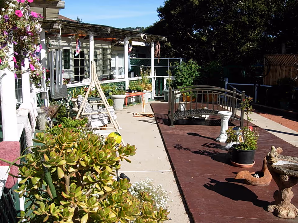 Outdoor patio area at Flanders Court of Carmel featuring a small decorative bridge over a raised platform, various potted plants, garden decorations including a birdbath and a snail sculpture, a swing chair, and a covered porch with hanging flowers and seating.