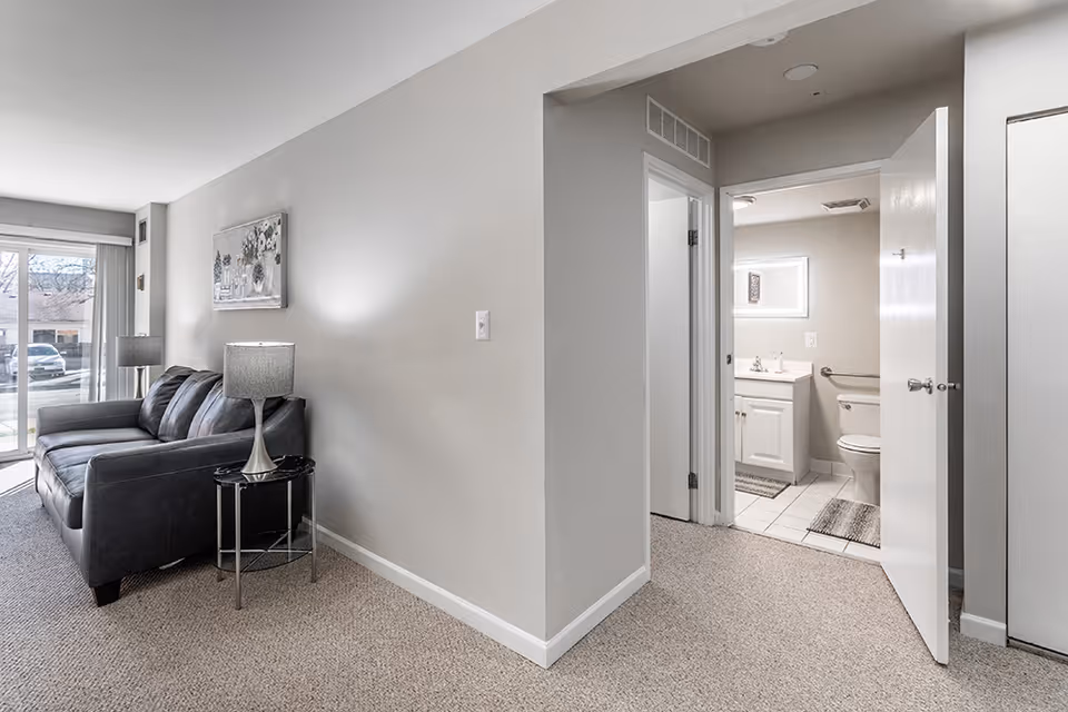 Interior view of a senior living facility showing a small living room with a dark gray sofa, a side table with a lamp, and a large window with vertical blinds. Adjacent to the living room is a hallway leading to a bathroom with a white vanity, toilet, and a mirror above the sink. The walls are painted light gray and the floor is carpeted except for the bathroom which has white tiles.