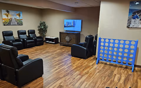 A senior living facility common area with several black recliner chairs arranged around a TV mounted on a wooden cabinet. The room has wood flooring, beige walls, a large blue and white Connect Four game, and framed pictures on the walls.