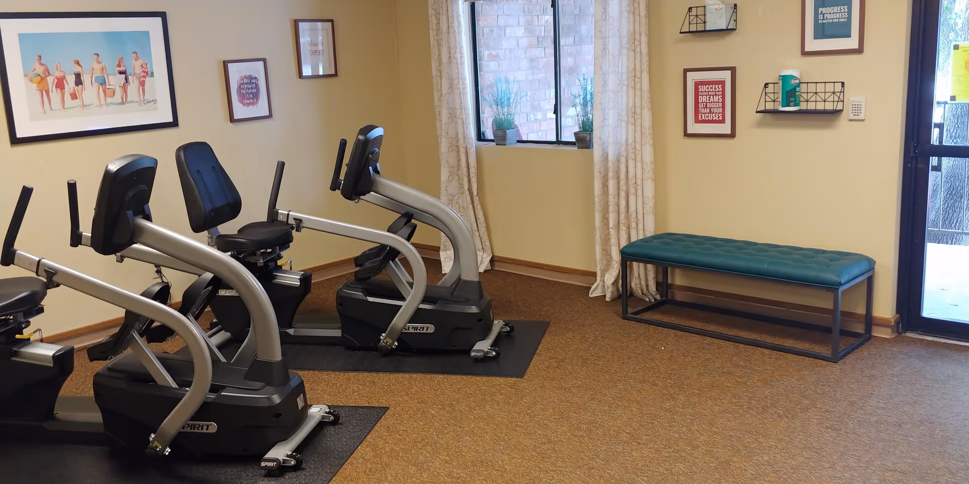 A small exercise room with three recumbent stationary bikes on black mats. The room has beige walls with framed artwork and motivational quotes. There is a window with curtains and a teal cushioned bench near a glass door.