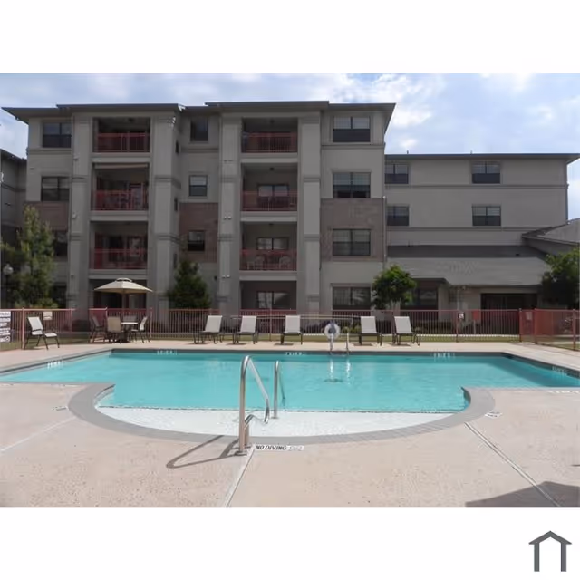 Outdoor swimming pool with clear blue water in front of a multi-story residential building with balconies. Poolside has lounge chairs and a table with an umbrella. The area is fenced with a red railing.