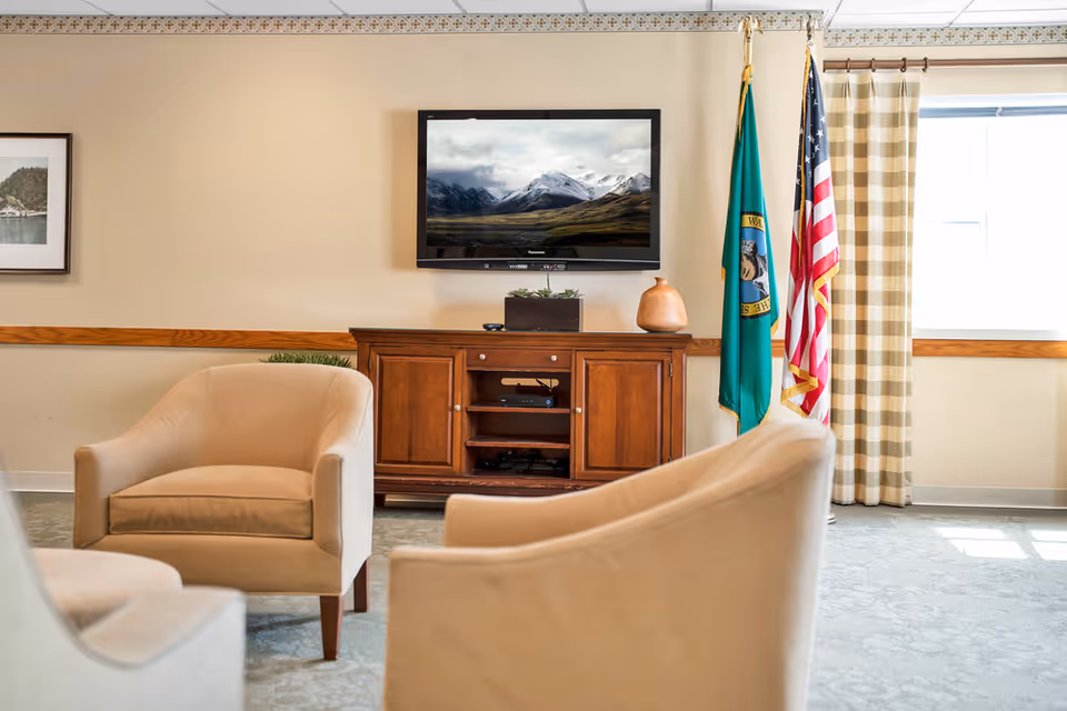 A cozy sitting area with beige armchairs arranged around a wooden cabinet with a flat-screen TV mounted above it. The TV displays a scenic mountain landscape. To the right of the cabinet are two flags, one of the United States and the other likely a state flag, next to a window with beige and white checkered curtains. The room has light-colored walls and carpeted flooring.