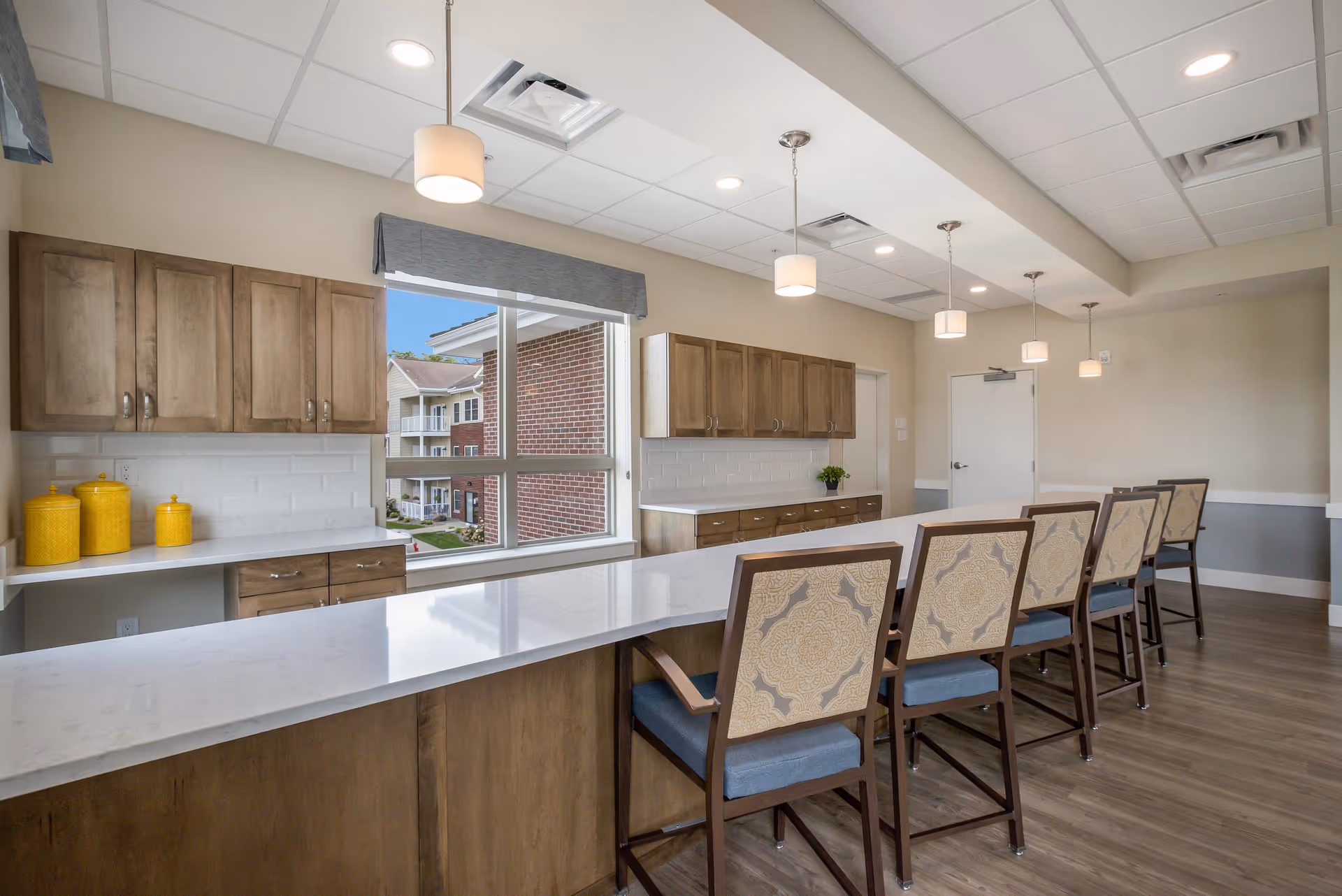 A modern kitchen area with a long white countertop island and six high-back chairs with patterned upholstery. The kitchen features wooden cabinets, white subway tile backsplash, and three pendant lights hanging from the ceiling. A large window shows an exterior view of a brick building and greenery outside.