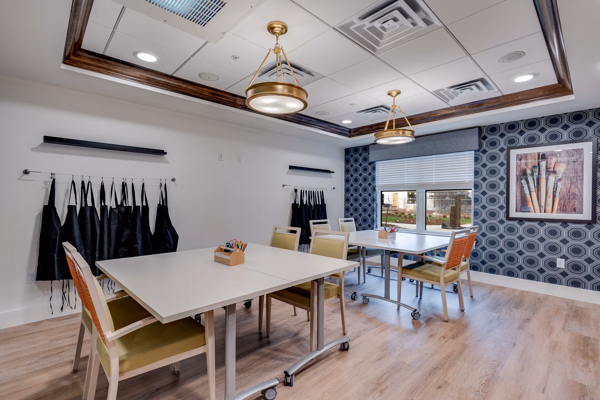 A bright, clean activity room with two white tables on wheels, surrounded by chairs with green cushions. Black aprons hang on racks along the white wall. The room features a large window with a view of outdoor greenery, a patterned blue wallpaper on one wall, and a framed picture of paintbrushes. Two modern pendant lights hang from a ceiling with recessed lighting and wooden trim.