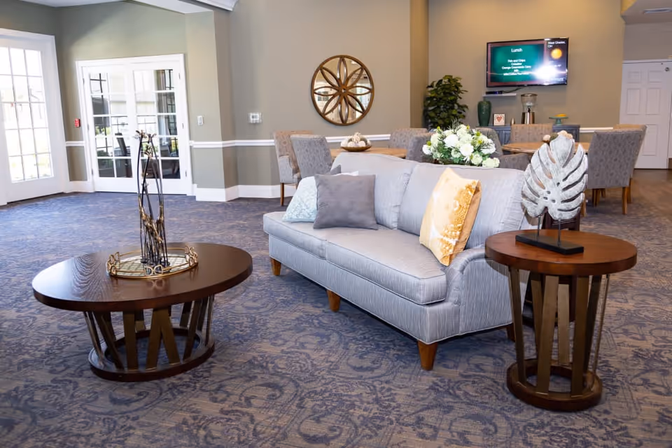 A cozy living room area with a light gray sofa adorned with three pillows in gray, blue, and yellow. In front of the sofa is a round wooden coffee table with a decorative centerpiece. To the right of the sofa is a matching round side table with a leaf-shaped sculpture. Behind the sofa, there is a dining table with six upholstered chairs. A round wall mirror and a flat-screen TV mounted on the wall are visible. The room has patterned blue carpet and light-colored walls with white trim. Large windows and glass doors allow natural light to enter the space.