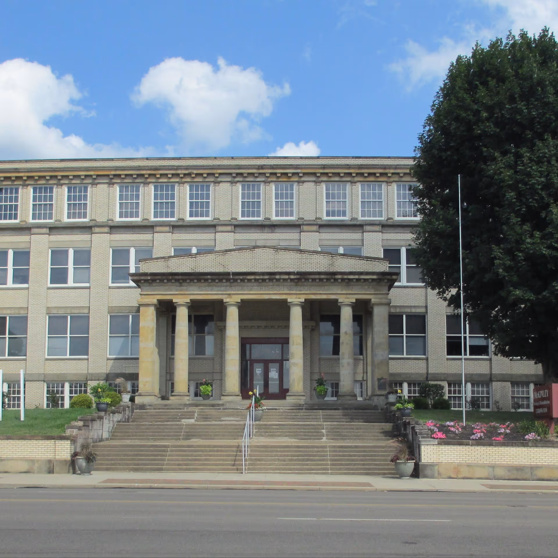 Front exterior view of McKinley Health Care Center, a large multi-story building with a wide staircase leading up to a covered entrance supported by six columns. There are flower pots and landscaping on either side of the stairs, a large tree to the right, and a clear blue sky with some clouds above.