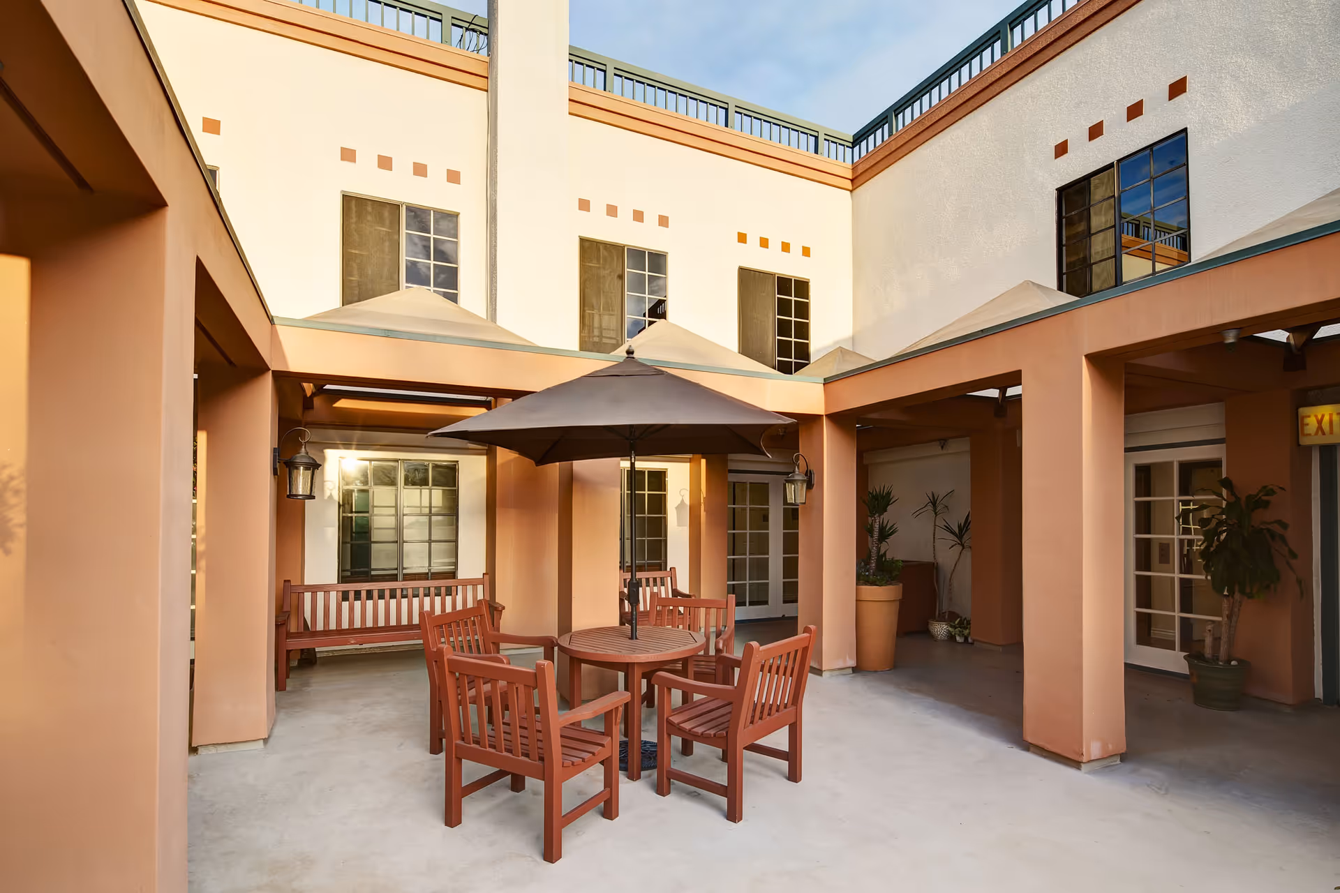 Outdoor courtyard area with a round wooden table and four wooden chairs under a large black umbrella. The courtyard is surrounded by peach-colored walls with windows and potted plants, and there is a wooden bench against one wall.