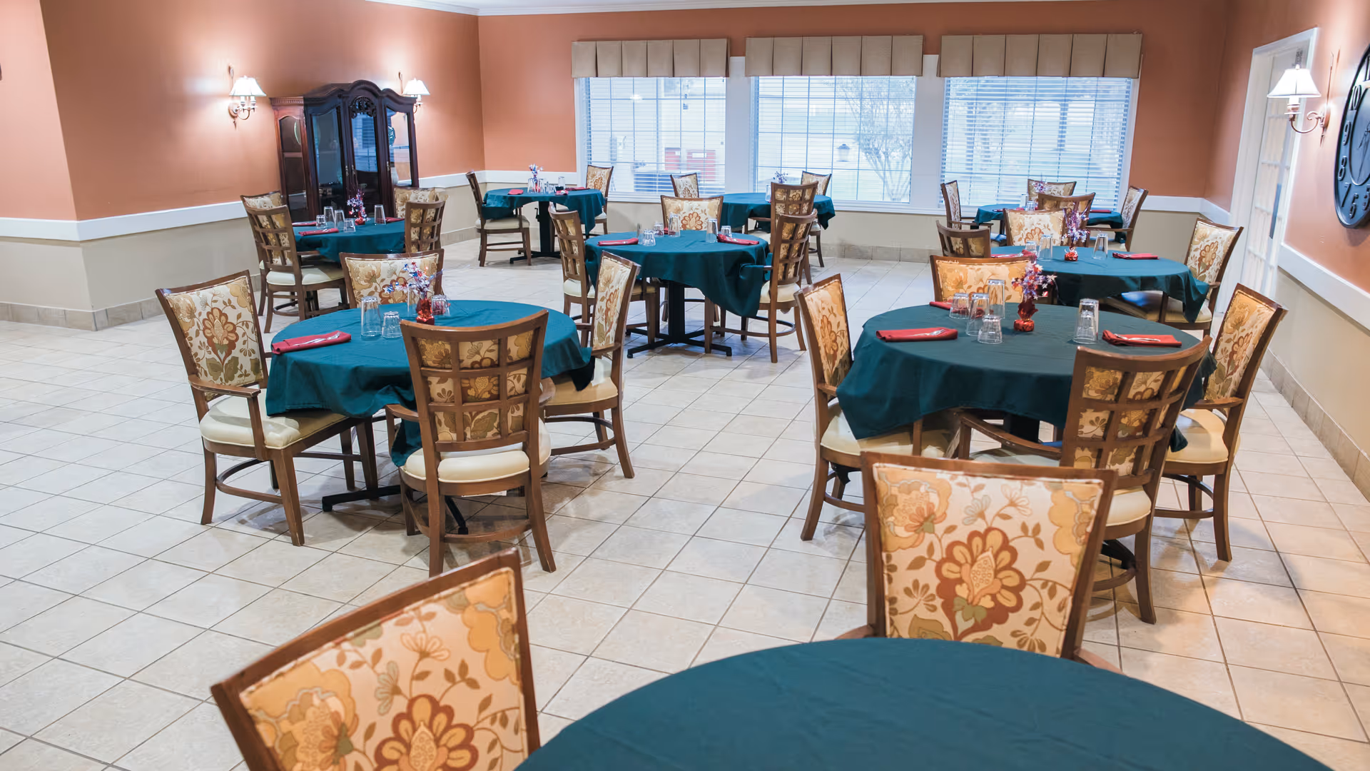 A dining room with multiple round tables covered with dark green tablecloths, each set with glasses and red napkins. The chairs have floral patterned upholstery. The room has tiled floors, peach-colored walls with white trim, and large windows letting in natural light.