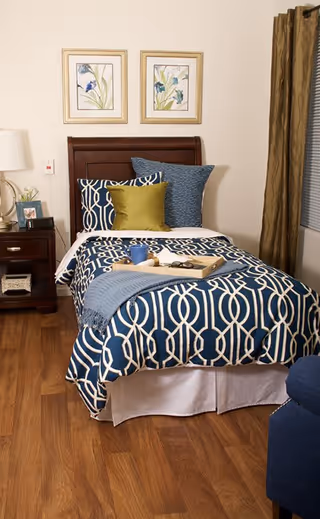 A neatly made single bed with a dark wooden headboard, decorated with blue and white patterned bedding, a green pillow, and a blue throw blanket. There is a wooden nightstand with a lamp, a framed photo, and a small plant next to the bed. Two framed floral artworks hang on the wall above the headboard. Brown curtains cover a window on the right side of the room, and the floor is wooden.