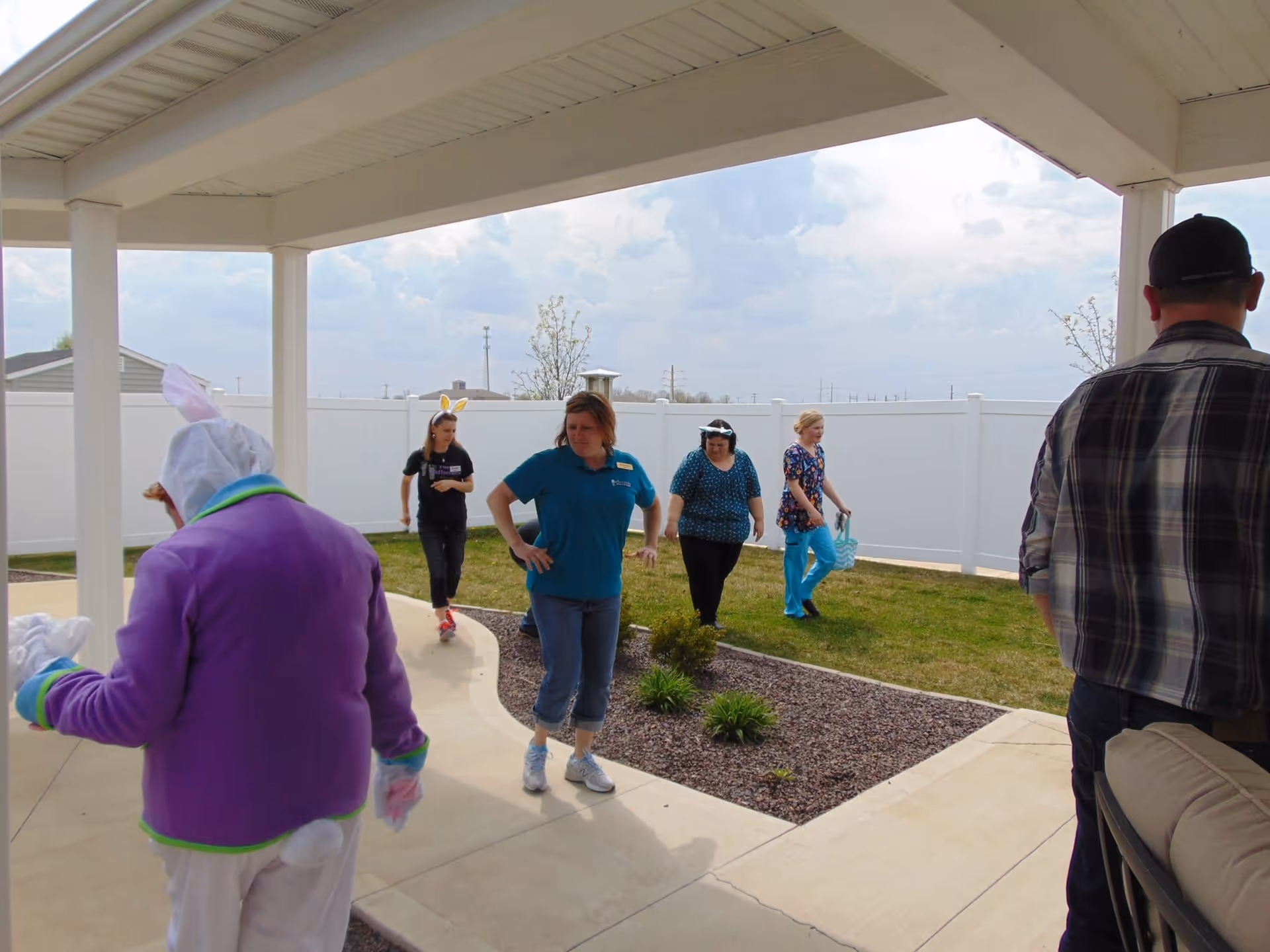 A group of people outdoors in a fenced yard area with a covered patio. One person is dressed in a bunny costume, and others are casually dressed, some wearing bunny ears. The sky is partly cloudy.