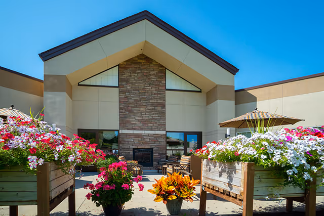 Outdoor courtyard with colorful flower planters, patio seating and umbrellas in front of a stone-accented building entrance under a clear blue sky.
