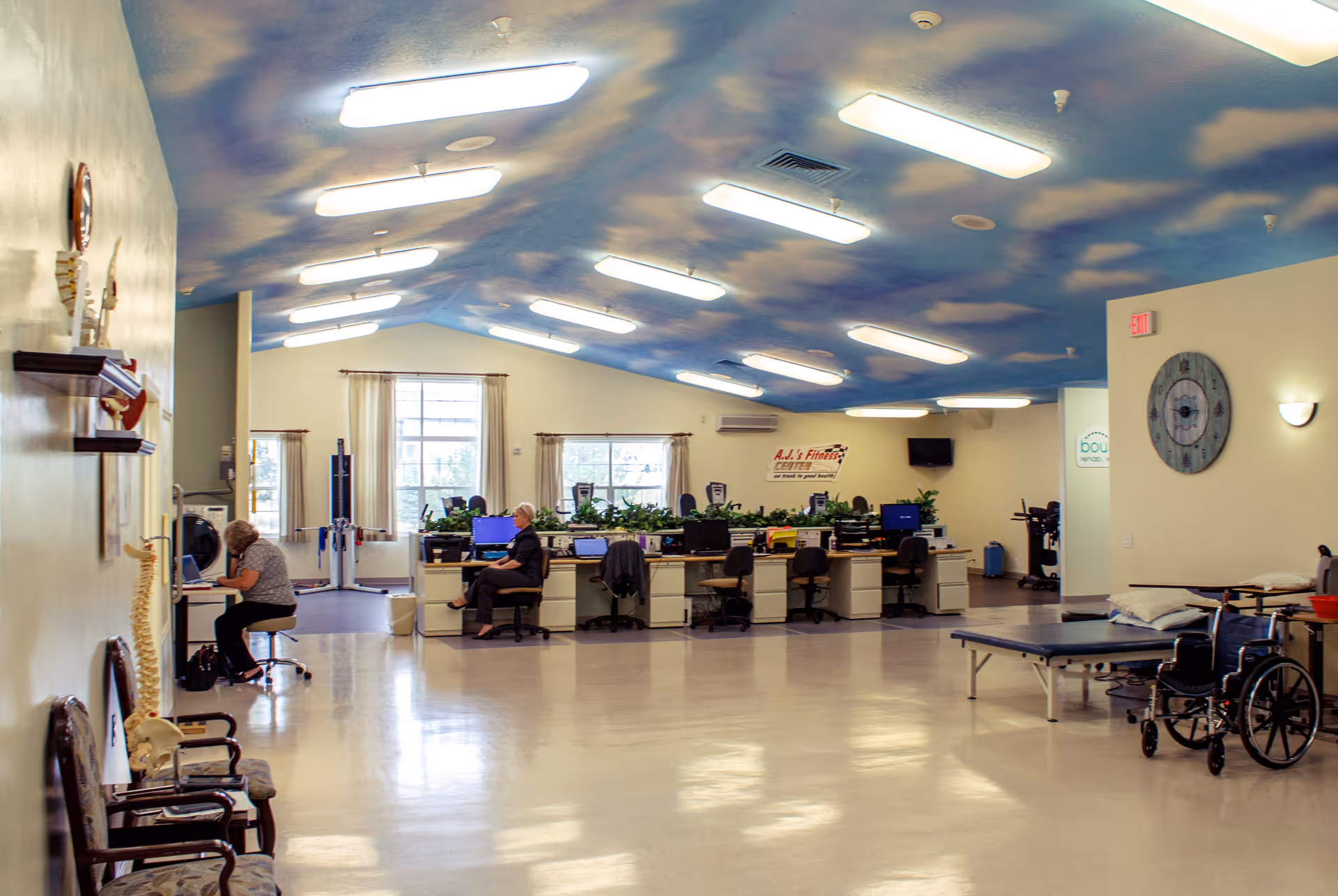 Large bright interior room with a cloud-painted ceiling, rows of desks with staff at computers, and therapy equipment including a treatment table and wheelchair.