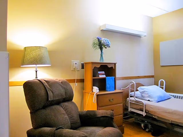 A cozy room with a brown recliner chair, a wooden nightstand with a vase of flowers and a blue gift bag, and a hospital-style bed with white railings and a pillow. The room has soft yellow walls and a lamp providing warm light.