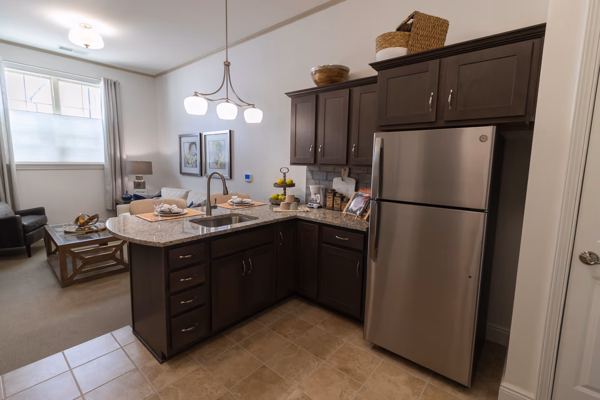 A modern kitchen area with dark wood cabinets, a stainless steel refrigerator, and a granite countertop island with a built-in sink. The kitchen opens into a living room with a sofa, armchair, coffee table, and two framed pictures on the wall. A three-light chandelier hangs above the kitchen island, and a window with curtains allows natural light into the space.