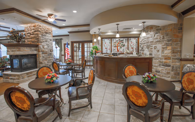 A cozy common area in a senior living facility featuring round tables with floral centerpieces and upholstered chairs with orange patterned backs. There is a stone fireplace on the left side, an American flag near the windows, and a curved wooden reception desk with pendant lights hanging above. The walls are decorated with stone accents and a stained glass window behind the desk.