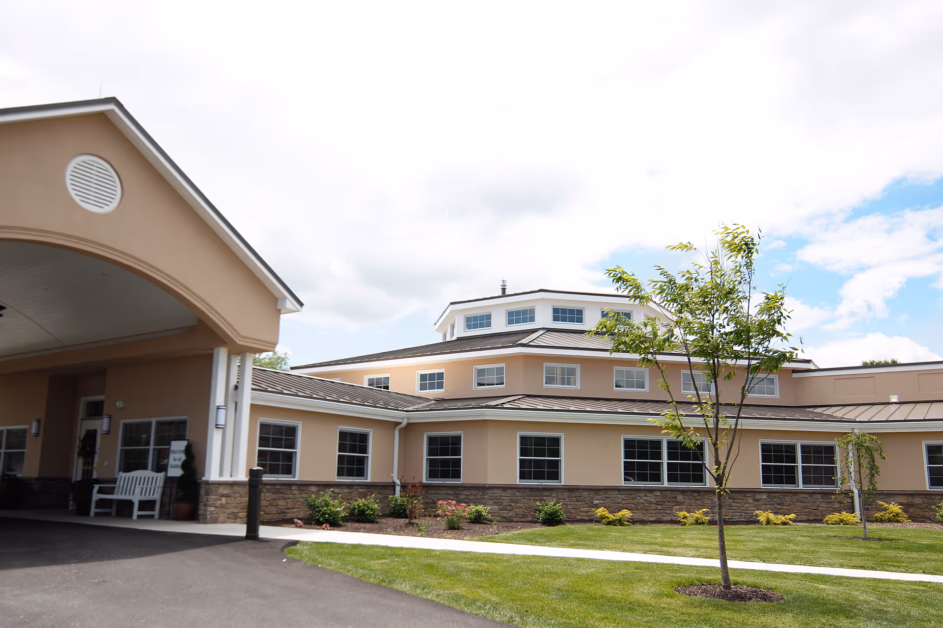 Exterior view of a beige and stone retirement community building with multiple windows, a covered entrance with a white bench, a small tree, and a well-maintained lawn under a partly cloudy sky.