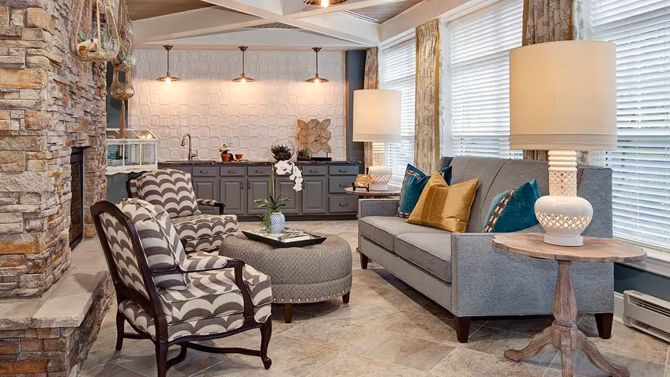 A cozy living room area featuring a gray sofa with colorful throw pillows, two patterned armchairs, a round ottoman with a tray and a small plant, a stone fireplace on the left, and a side table with a white lamp. The background shows a wall with gray cabinetry, a sink, and three pendant lights above. Large windows with blinds allow natural light to fill the space.