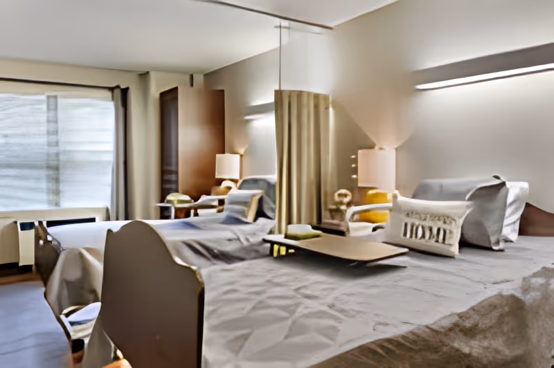 A well-lit room with two hospital-style beds separated by a privacy curtain. Each bed has pillows and a small bedside table with a lamp. There is a large window with curtains on the left side of the room, and the overall decor is simple and functional.