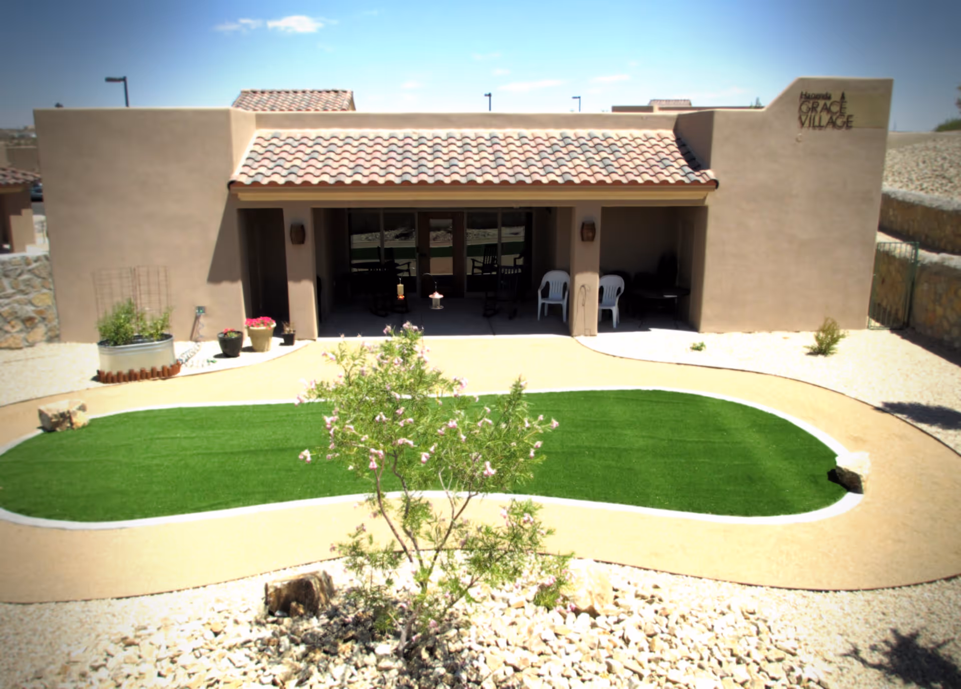 Exterior view of a single-story building with a tiled roof and beige stucco walls at Haciendas at Grace Village. In front of the building is a small landscaped area with a green artificial turf patch surrounded by a beige pathway and desert rocks. There are potted plants near the entrance and two white plastic chairs under a covered patio.