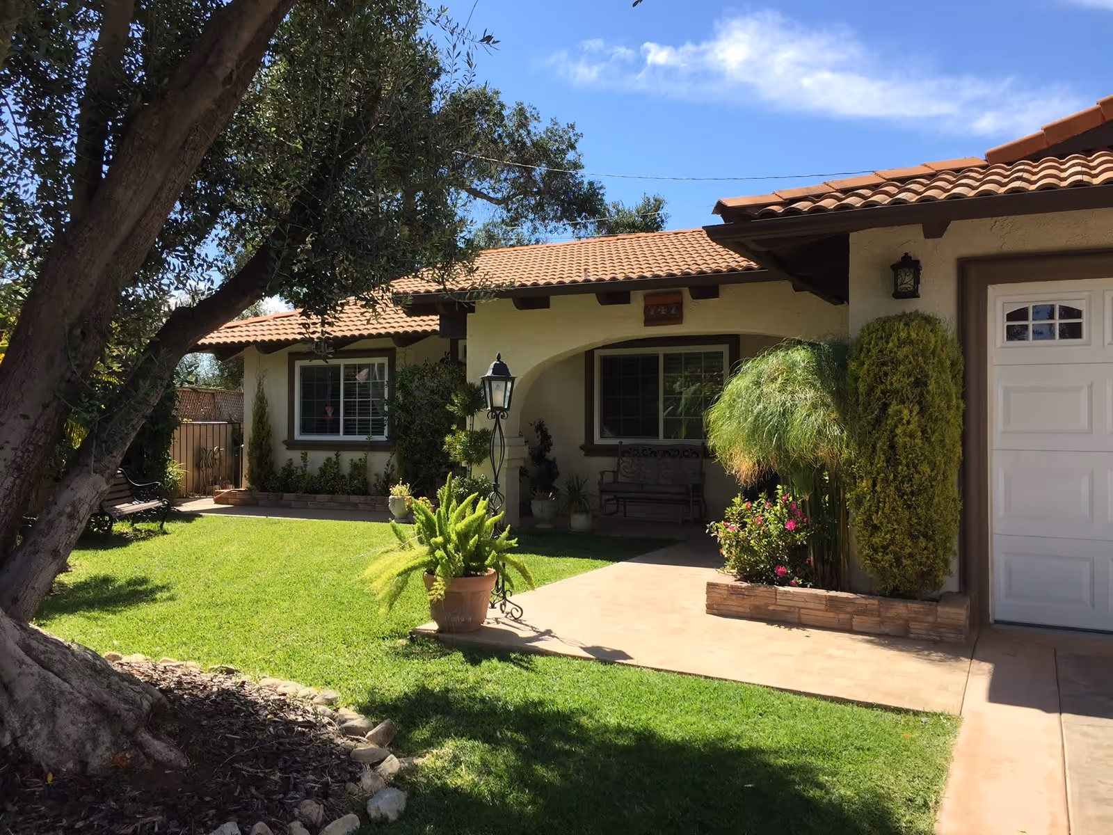 Front exterior view of a single-story house with a tiled roof, beige walls, and brown trim. The house has two windows with white shutters, a white garage door, and a small covered porch with a bench. The front yard features a well-maintained green lawn, various potted plants, shrubs, a tree, and a black metal lamp post. The sky is clear and blue.