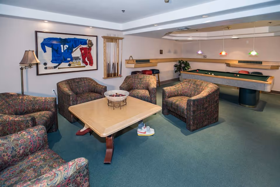 A furnished game room with upholstered chairs around a coffee table and a pool table in the background.