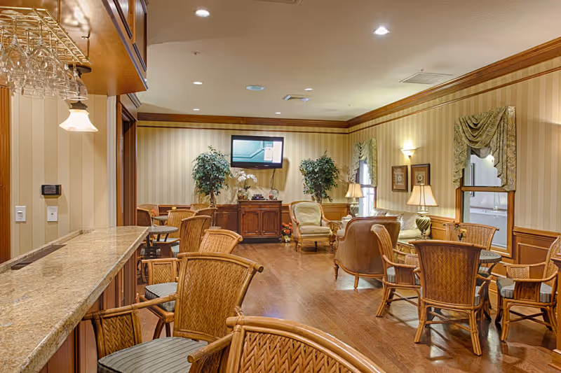 A warm, well-lit common room with wicker chairs and tables, a granite-topped bar, upholstered armchairs, potted plants, and a wall-mounted TV.