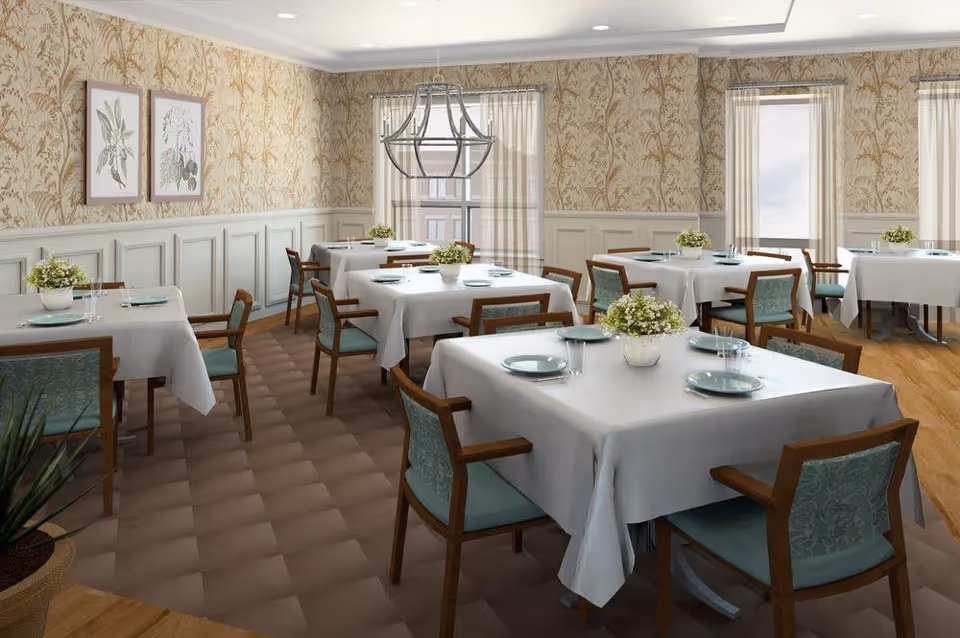 A dining room with several square tables covered with white tablecloths, each set with plates, glasses, and small flower arrangements. The room has patterned wallpaper, wainscoting, large windows with sheer curtains, and a modern chandelier hanging from the ceiling. Wooden chairs with teal upholstered seats and backs surround the tables.