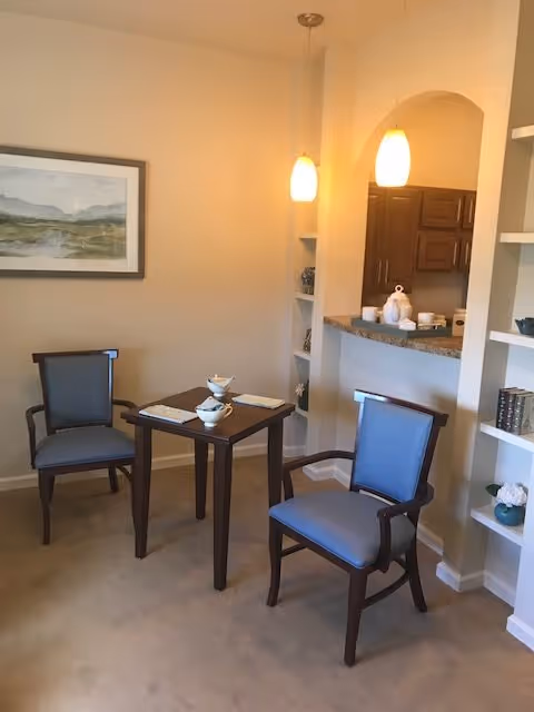 A small dining area with two wooden chairs with blue cushions around a square wooden table set with two bowls and placemats. The area is adjacent to a kitchen counter with two hanging pendant lights above. There is a framed landscape painting on the wall and built-in shelves with decorative items.