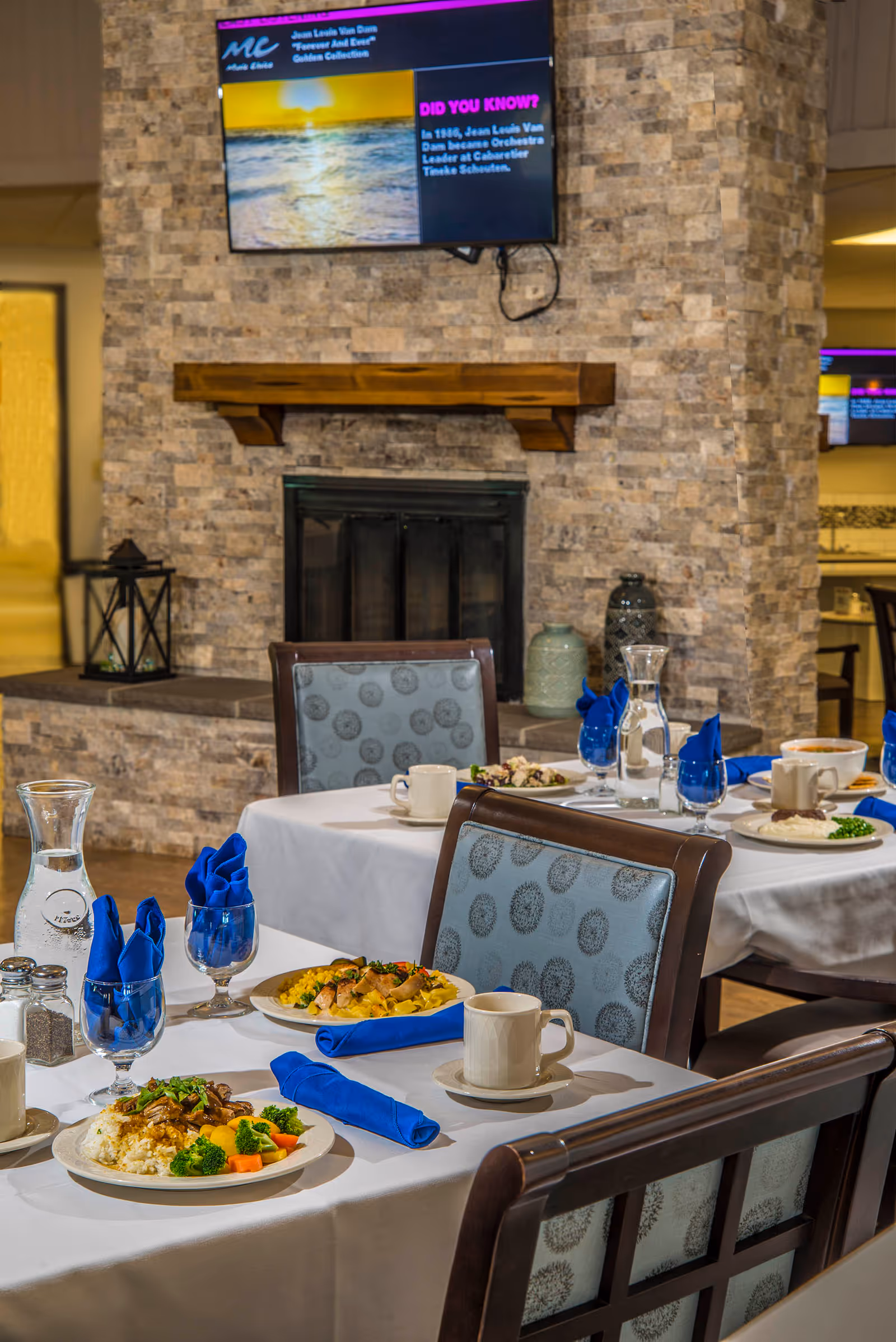 Dining room with neatly set tables featuring plated meals and blue napkins in front of a stone fireplace with a TV above it.