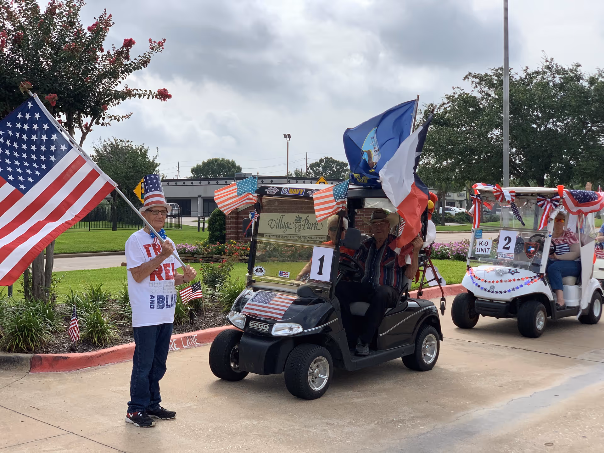 An elderly man wearing a patriotic hat and a red, white, and blue shirt stands holding a large American flag next to a golf cart decorated with American flags and other flags. Another golf cart decorated with patriotic bunting and a sign reading 'K-9 UNIT' is visible behind them. The scene is outdoors near the entrance sign of Village on the Park Steeplechase, with trees and a cloudy sky in the background.
